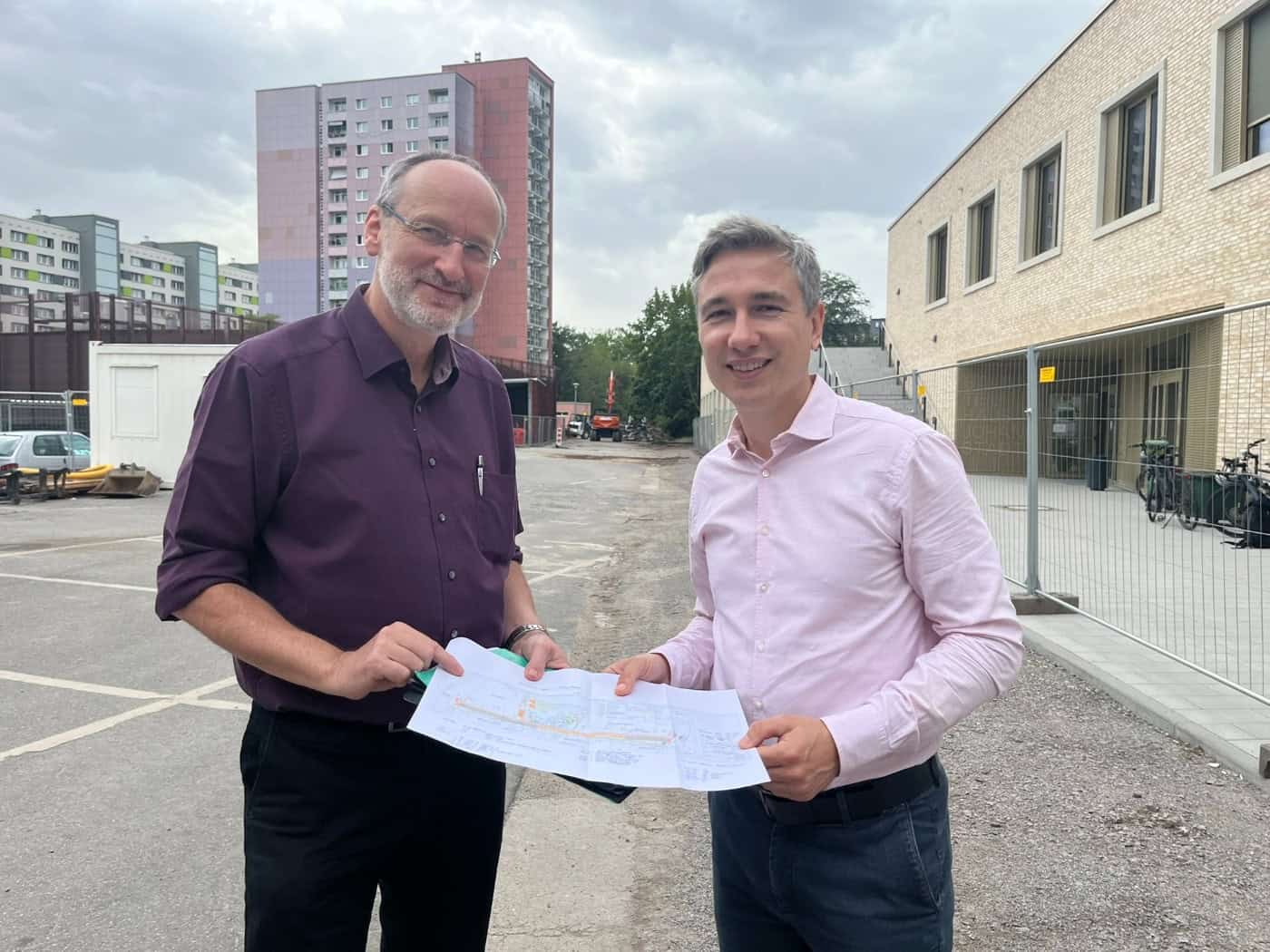 Stephan Kühn in hellem Hemd und Thomas Pieper im lila Hemd halten gemeinsam einen Bauplan vor einer Baustelle am Stadtteilhaus in Dresden-Johannstadt. Im Hintergrund sind Wohnblöcke und ein Bauzaun zu sehen.