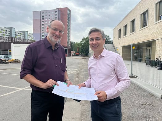 Stephan Kühn in hellem Hemd und Thomas Pieper im lila Hemd halten gemeinsam einen Bauplan vor einer Baustelle am Stadtteilhaus in Dresden-Johannstadt. Im Hintergrund sind Wohnblöcke und ein Bauzaun zu sehen.