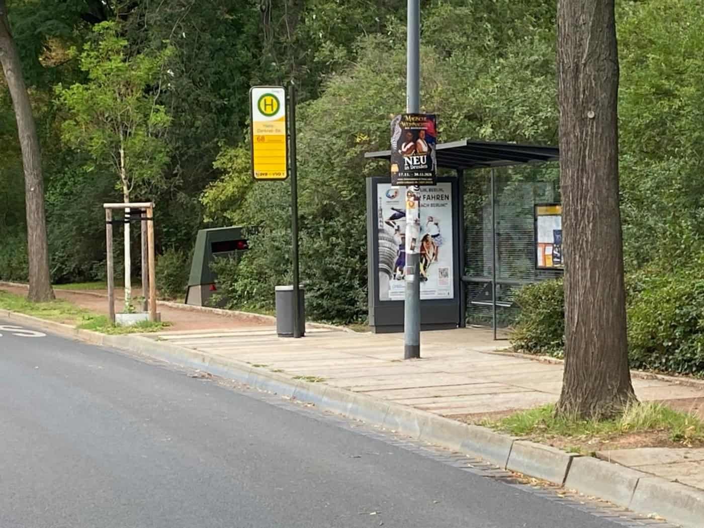 Blick auf eine Straße an einer Bushaltestelle in Dresden mit dichtem Gebüsch und Bäumen im Hintergrund. Rechts neben dem gläsernen Wartehäuschen duckt sich ein olivgrüner, kastenartiger Messanhänger (Superblitzer) ins Grün.
