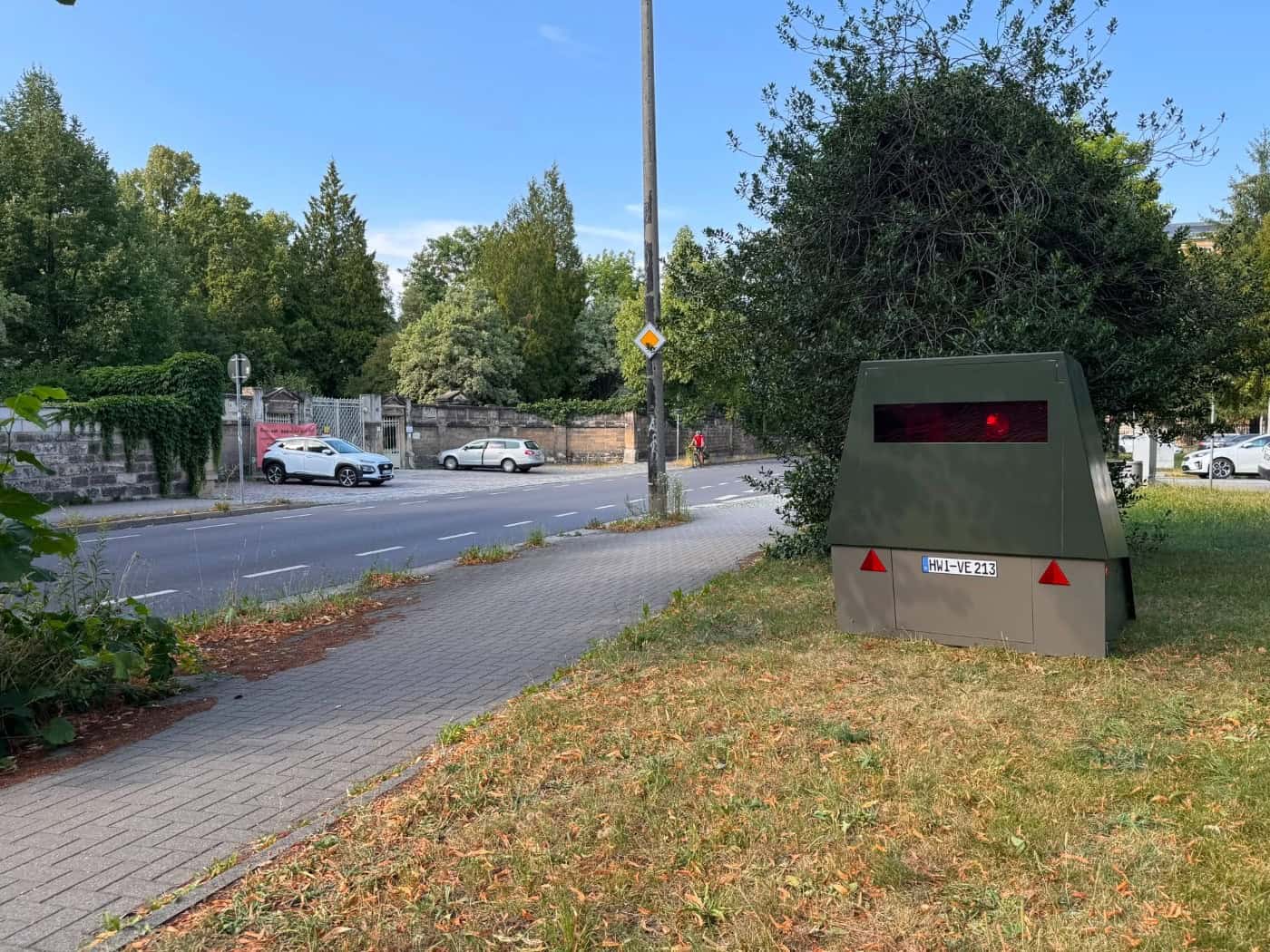 Ein olivgrüner Blitzeranhänger mit dem Kennzeichen HWI-VE 213 steht gut getarnt auf einer Grünfläche neben dem Gehweg an der Chemnitzer Straße in Dresden. Im Hintergrund verläuft die Fahrbahn mit geparkten Autos.