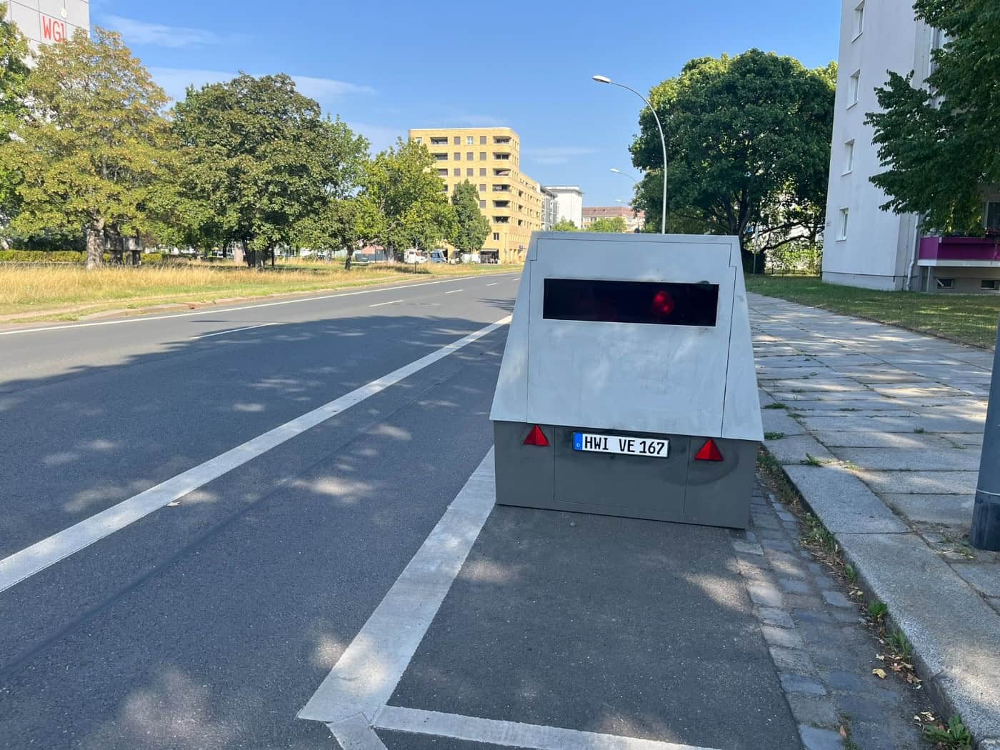 Ein grauer Blitzeranhänger mit dem Kennzeichen HWI-VE 167 steht am Straßenrand der Striesener Straße in Dresden. Im Hintergrund verläuft die asphaltierte Straße vorbei an Wohngebäuden und Bäumen.