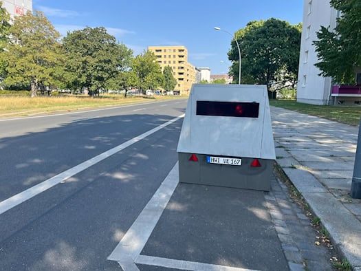 Ein grauer Blitzeranhänger mit dem Kennzeichen HWI-VE 167 steht am Straßenrand der Striesener Straße in Dresden. Im Hintergrund verläuft die asphaltierte Straße vorbei an Wohngebäuden und Bäumen.