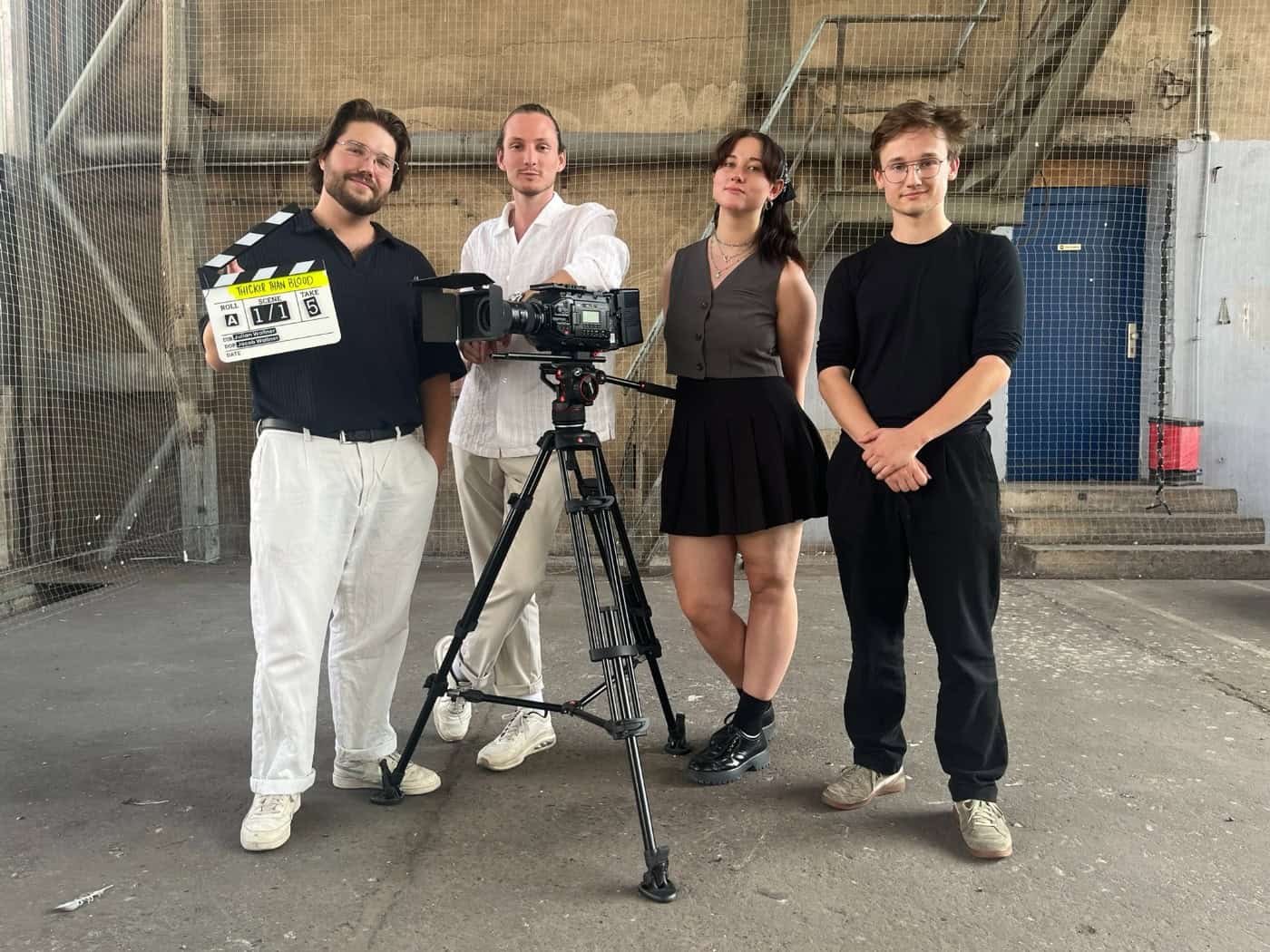 Vier junge Filmschaffende stehen in einer Industriehalle hinter einer Kamera auf einem Stativ. Ganz links hält Julian Wallner eine Filmklappe mit der Aufschrift „Thicker Than Blood“. Neben ihm stehen Theodor Glöer, Eleanor Großhennig und Jacob Wallner.