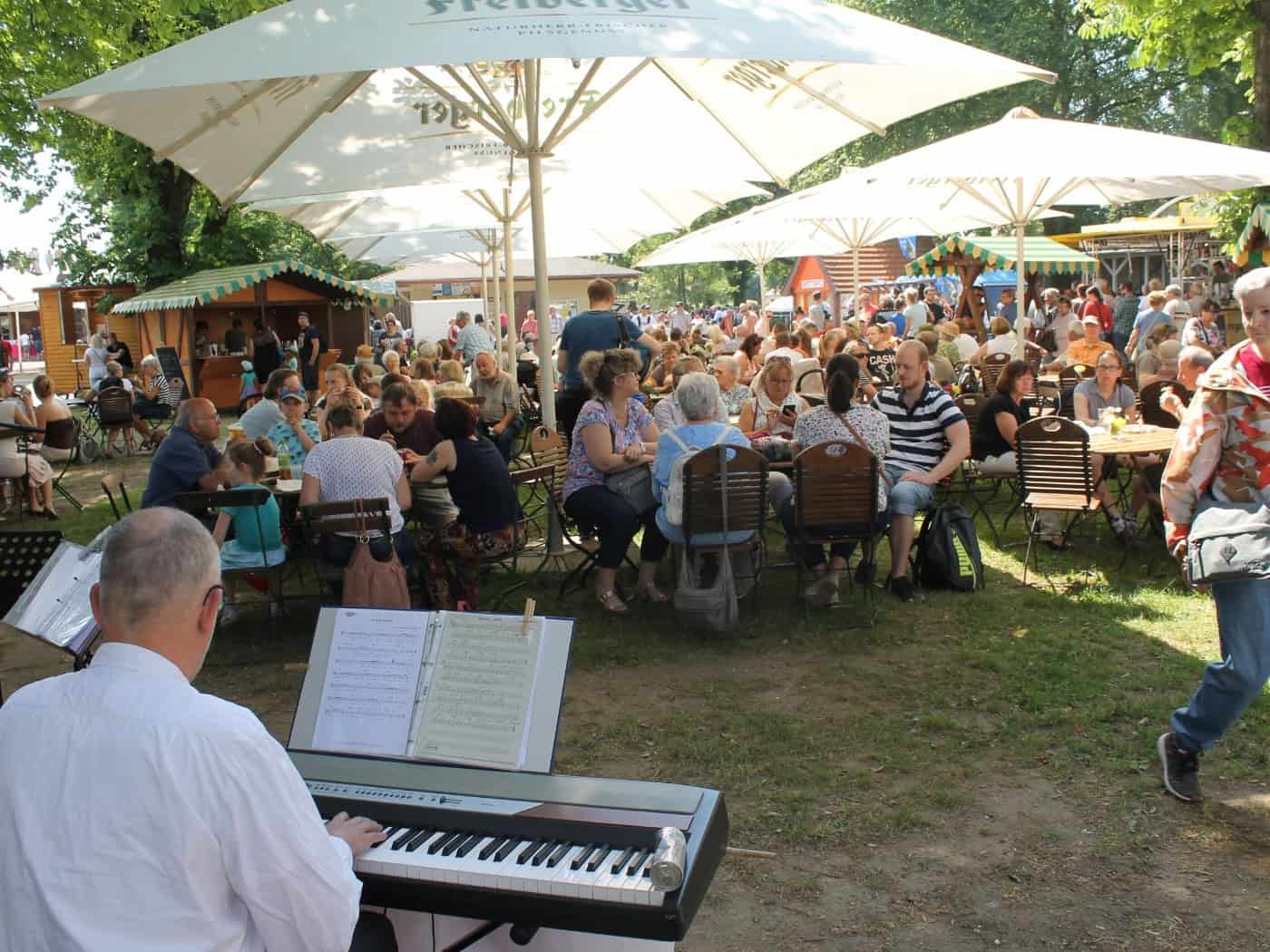 Blick von hinten auf einen Keyboardspieler in einem voll besetzten Biergarten unter Sonnenschirmen. Im Hintergrund schlendern Menschen über einen Markt mit Holzbuden auf dem schattigen Gelände der Galopprennbahn Dresden.