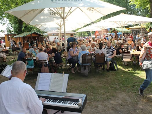 Blick von hinten auf einen Keyboardspieler in einem voll besetzten Biergarten unter Sonnenschirmen. Im Hintergrund schlendern Menschen über einen Markt mit Holzbuden auf dem schattigen Gelände der Galopprennbahn Dresden.