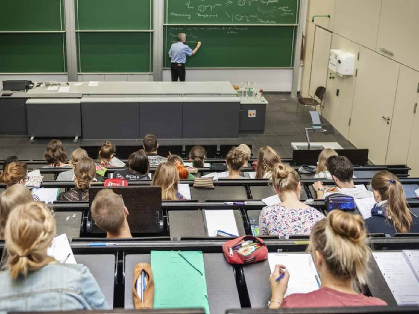 Blick von hinten auf Studierende, die in den Sitzreihen eines Hörsaals sitzen und mitschreiben. Vorne an einer großen grünen Tafel schreibt ein Dozent chemische Formeln an.