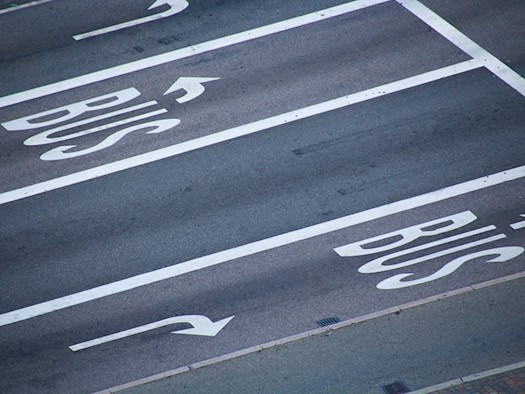 Draufsicht auf eine asphaltierte Straße mit weißen Fahrbahnmarkierungen für zwei Busspuren. Das Wort "BUS" ist in großen Buchstaben auf den Asphalt gemalt, jeweils mit einem Pfeil nach links und rechts.