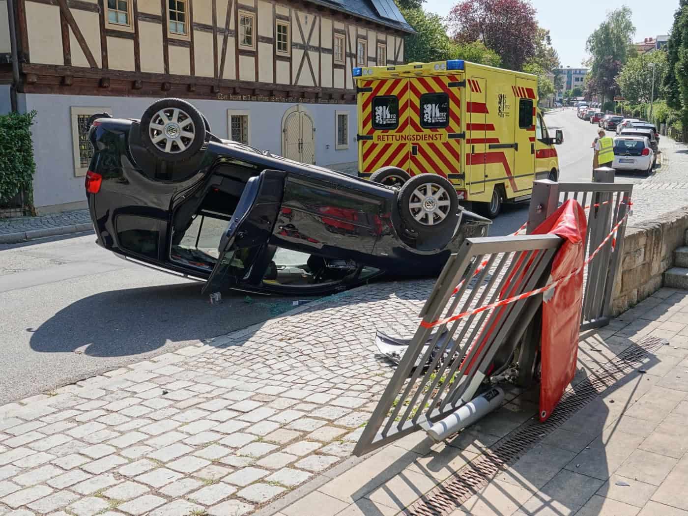 Ein schwarzer VW Touran liegt auf dem Dach auf einem Gehweg an einer beschädigten Toranlage. Im Hintergrund steht ein gelb-roter Rettungswagen der Dresdner Feuerwehr vor einem Fachwerkhaus.