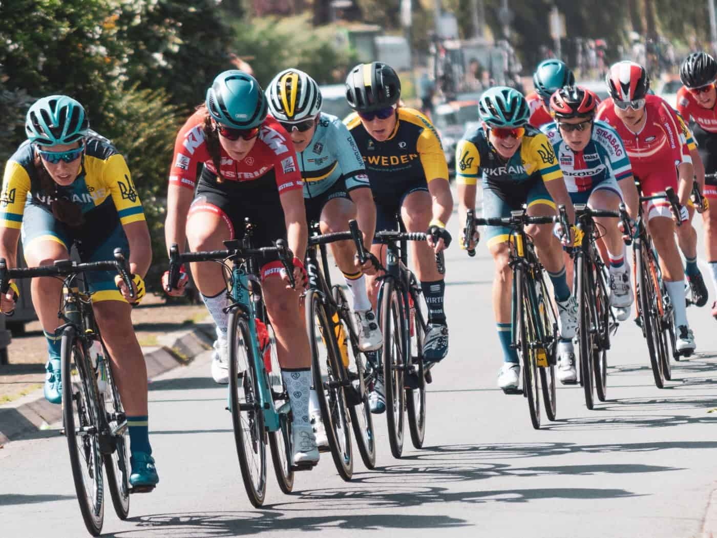 Eine Gruppe von Radrennfahrerinnen in bunten Trikots und Helmen fährt während eines Rennens dicht hintereinander auf einer asphaltierten Straße im Sonnenschein.