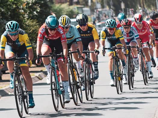 Eine Gruppe von Radrennfahrerinnen in bunten Trikots und Helmen fährt während eines Rennens dicht hintereinander auf einer asphaltierten Straße im Sonnenschein.