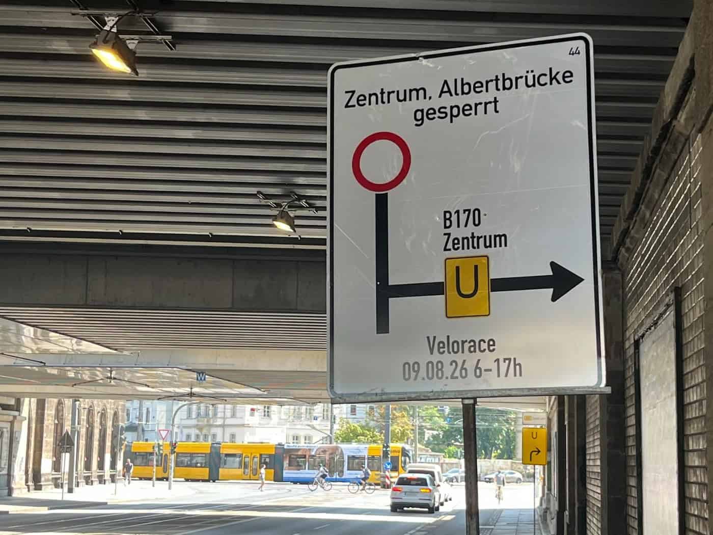 Ein weißes Umleitungsschild unter einer Brücke weist auf die Sperrung von Zentrum und Albertbrücke für das Velorace am 09.08.26 von 6 bis 17 Uhr hin. Im Hintergrund sind eine gelbe Straßenbahn und Autos auf einer sonnigen Straße zu sehen.