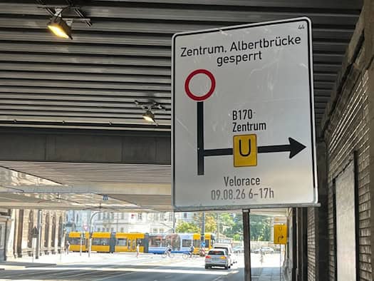 Ein weißes Umleitungsschild unter einer Brücke weist auf die Sperrung von Zentrum und Albertbrücke für das Velorace am 09.08.26 von 6 bis 17 Uhr hin. Im Hintergrund sind eine gelbe Straßenbahn und Autos auf einer sonnigen Straße zu sehen.