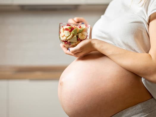 Nahaufnahme des unbedeckten Babybauchs einer schwangeren Frau in einer hellen Küche. Sie hält eine kleine Glas-Schale mit frischen Obststücken wie Bananen, Apfel und Granatapfelkernen in beiden Händen.