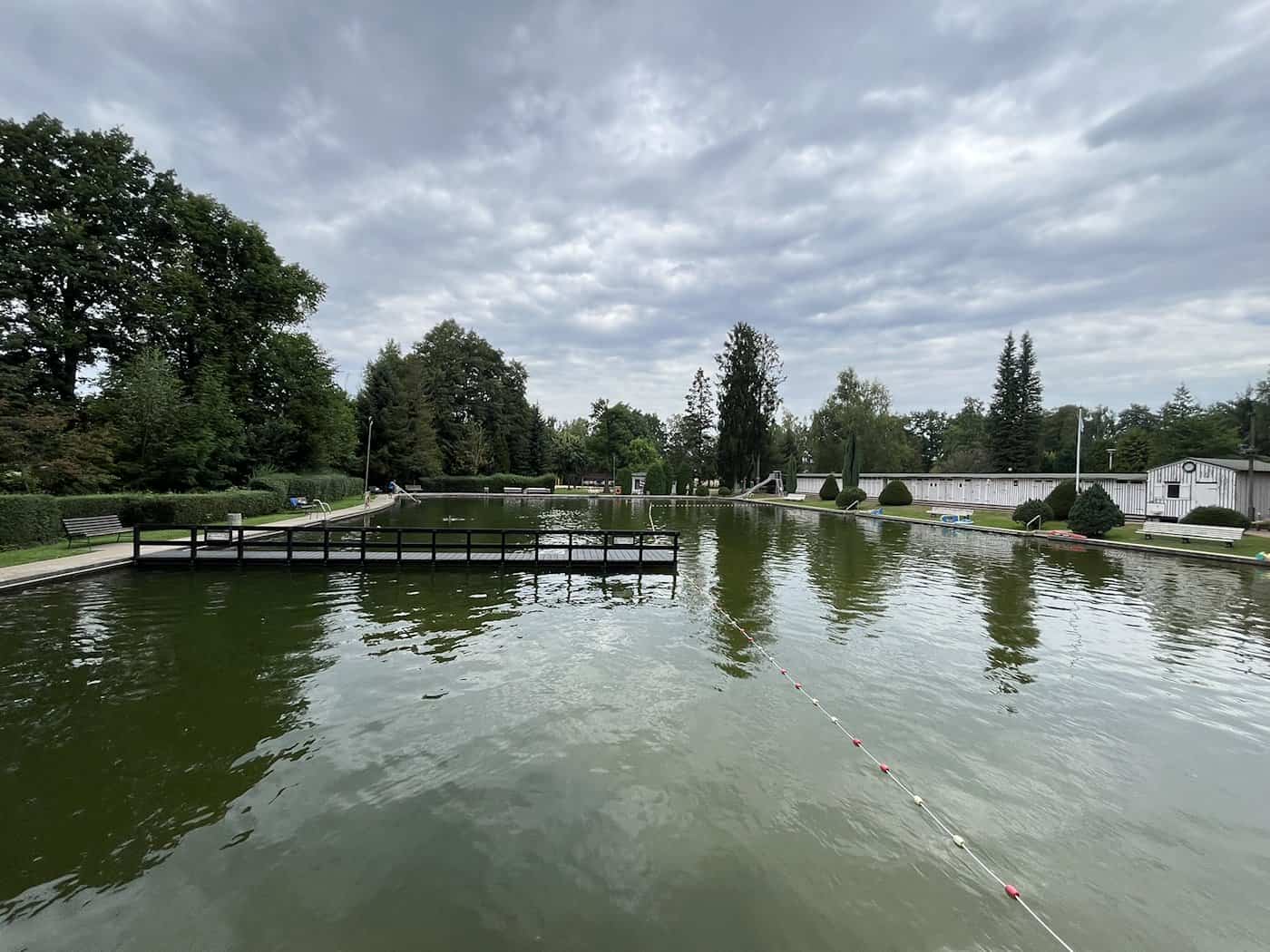 Panorama eines großen Naturbads mit grün schimmerndem Wasser, einem Holzsteg in der Mitte, grünen Bäumen am Ufer und weißen Umkleidekabinen unter bewölktem Himmel.