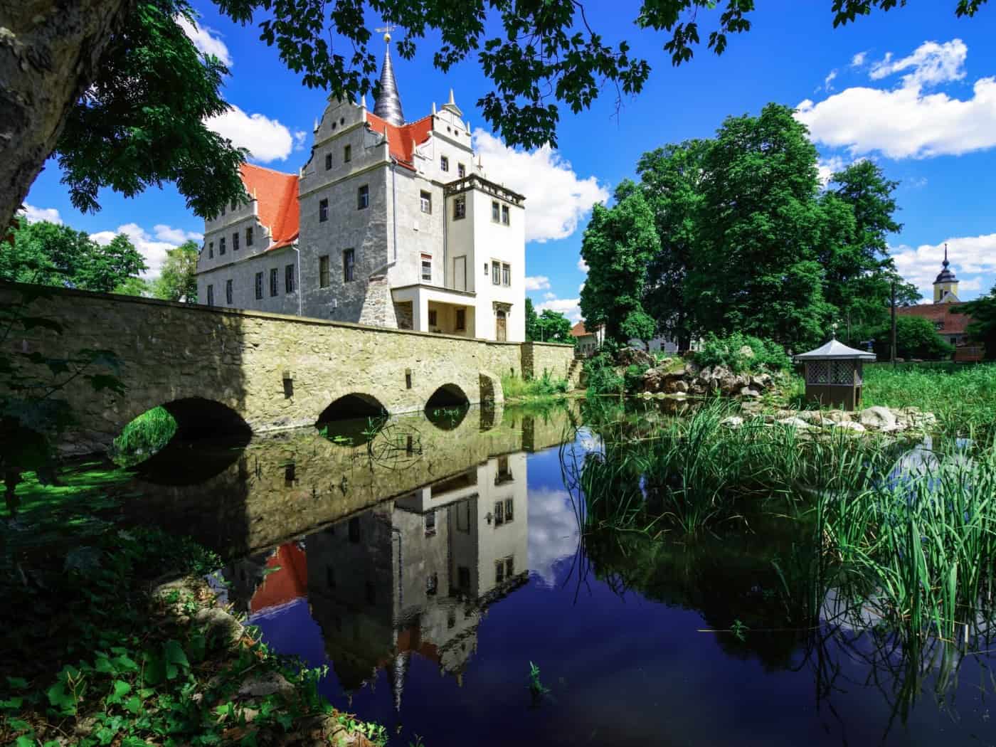 Das sanierte Wasserschloss Oberau spiegelt sich im Schlossteich hinter einer Steinbogenbrücke. Am Ufer wächst dichtes Schilf unter einem blauen Sommerhimmel mit weißen Wolken.