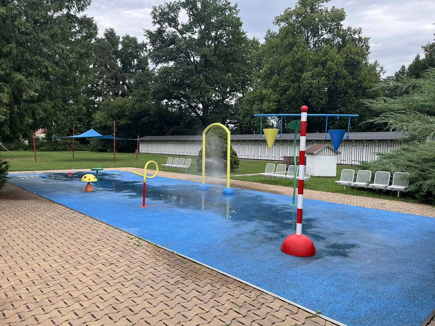Farbintensiver Wasserspielplatz im Freien mit blauem Antirutsch-Boden, roten und gelben Wasserduschen sowie bunten Eimern vor einer Wiese und Bäumen.