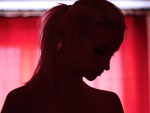 Symbolfoto: Nahaufnahme einer Frauensilhouette mit Pferdeschwanz im Gegenlicht. Der Blick ist gesenkt, im Hintergrund ist ein dunkelrot beleuchteter Vorhang zu sehen.