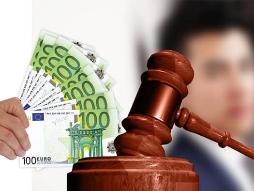Symbolfoto zum Thema Gerichtsurteil und Geldstrafe: Eine Hand hält gefächerte 100-Euro-Geldscheine neben einem hölzernen Richterhammer auf einem Sockel. Im unscharfen Hintergrund ist ein Mann zu erkennen.