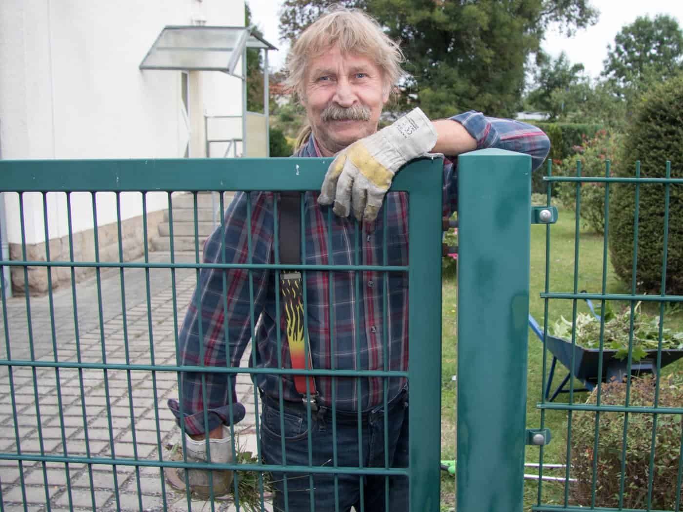 Ein lächelnder Mann mit Oberlippenbart, kariertem Hemd und Arbeitshandschuhen lehnt sich auf ein grünes Metalltor eines Gartenzauns vor einem Wohnhaus.