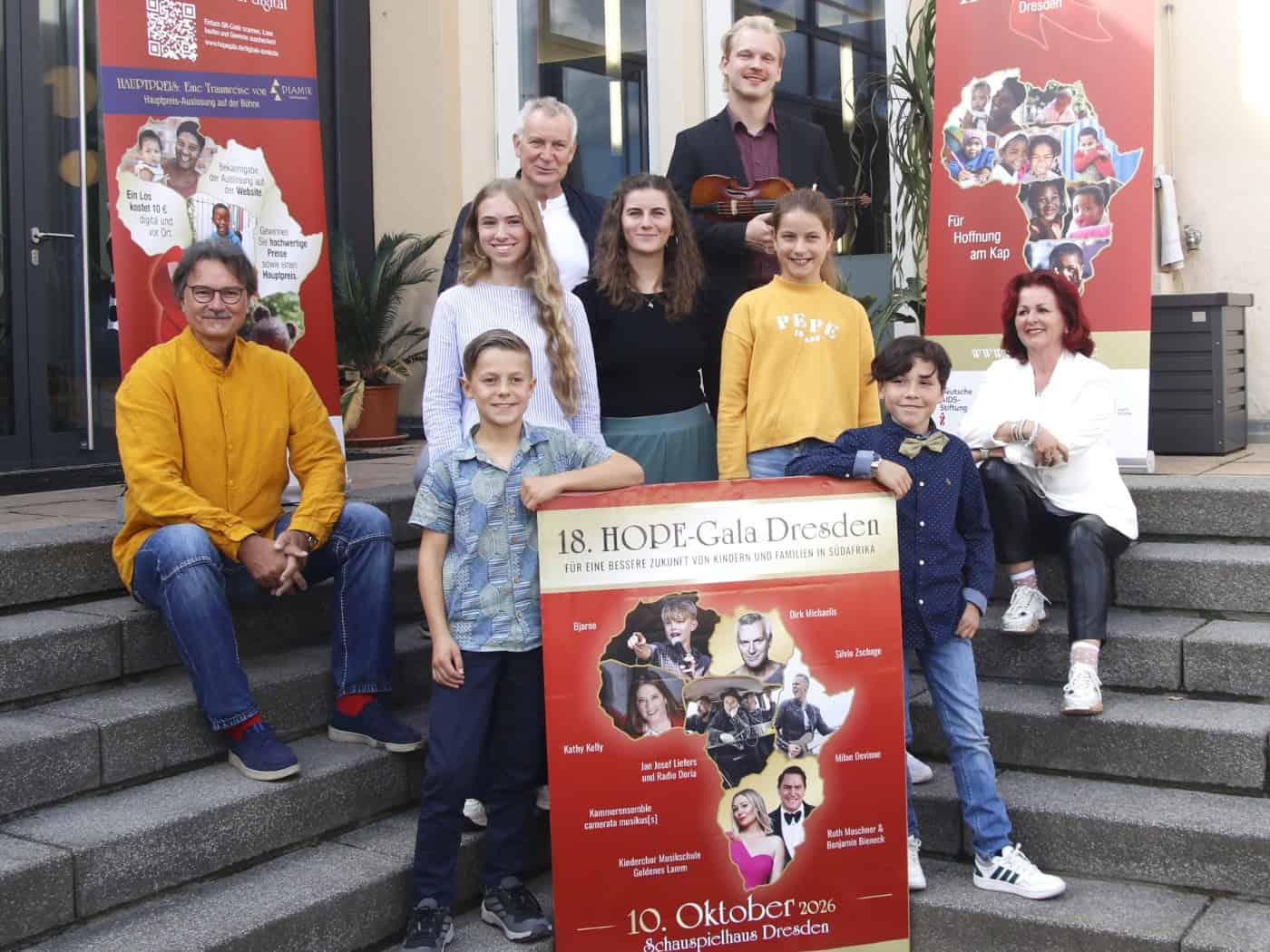 Eine Gruppe von Erwachsenen und Kindern posiert lächelnd auf einer Außentreppe. Zwei Jungen halten vorne ein rotes Plakat zur 18. HOPE-Gala Dresden. Rechts sitzt Initiatorin Viola Klein in Weiß, ganz links sitzt Prof. Milko Kersten im gelben Hemd auf einer Stufe.