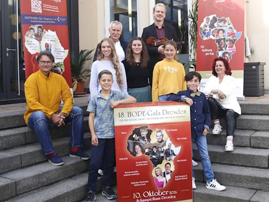 Eine Gruppe von Erwachsenen und Kindern posiert lächelnd auf einer Außentreppe. Zwei Jungen halten vorne ein rotes Plakat zur 18. HOPE-Gala Dresden. Rechts sitzt Initiatorin Viola Klein in Weiß, ganz links sitzt Prof. Milko Kersten im gelben Hemd auf einer Stufe.