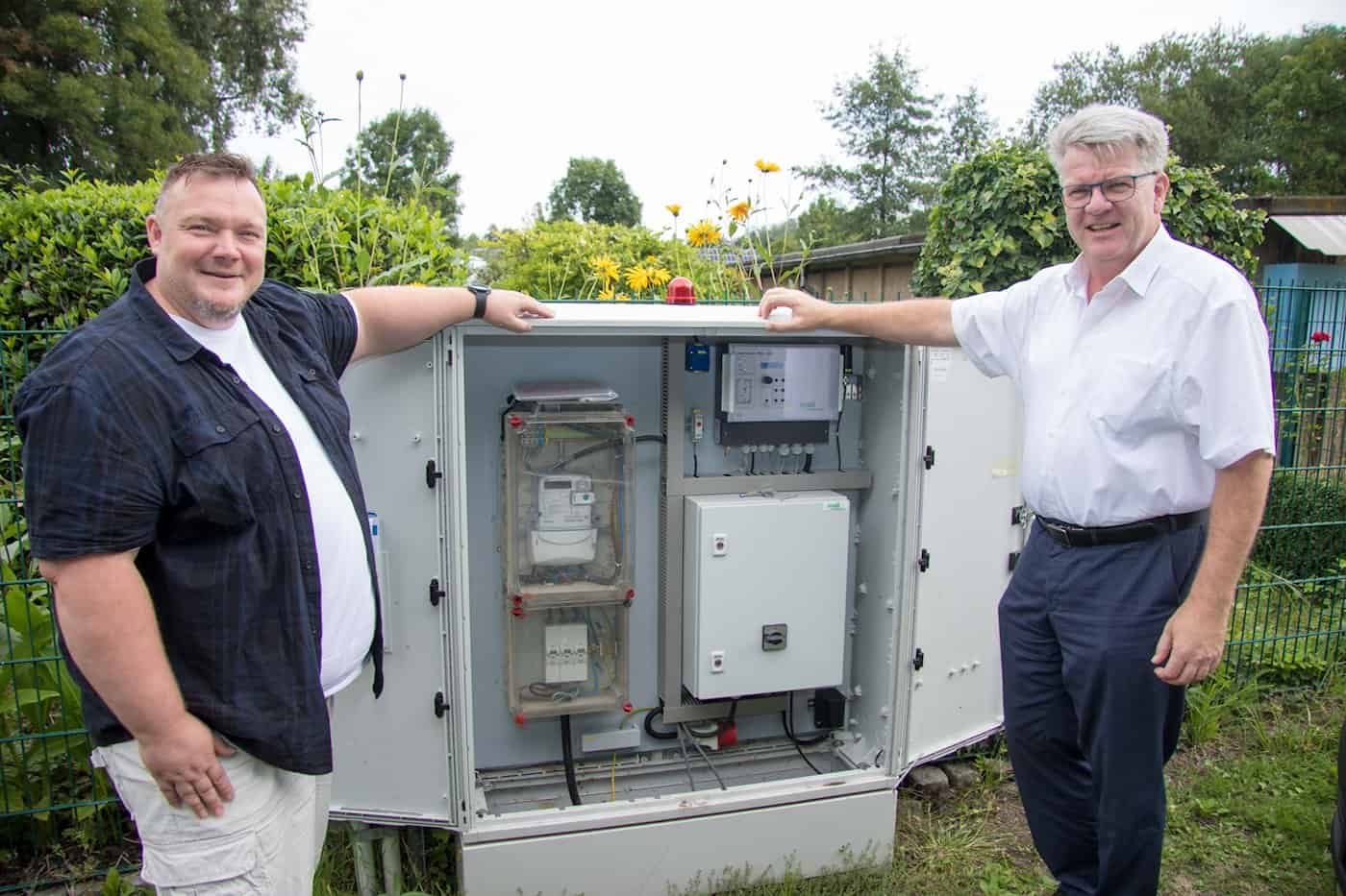 Zwei Männer stehen links und rechts neben einem großen, geöffneten Elektro-Schaltschrank im Freien, der Steuerungstechnik und Zähler für eine Abwasseranlage enthält.