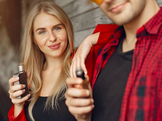 Eine Frau hält ein kompaktes Pod-System, ein Mann eine größere E-Zigarette mit Tank – Vergleich von Einweg-Vapes und Mehrweg-Systemen.