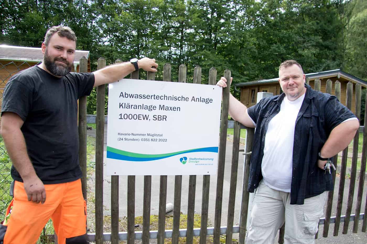 Zwei Mitarbeiter stehen an einem Holzzaun neben einem Schild mit der Aufschrift „Abwassertechnische Anlage Kläranlage Maxen 1000EW, SBR“ der Stadtentwässerung Dresden.