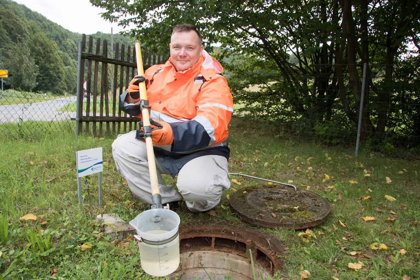 Ein Mann in orange-blauer Warnjacke kniet neben einem geöffneten Kanaldeckel und hält eine Schöpfstange mit einem transparenten Messbecher voller Wasser aus dem Schacht.