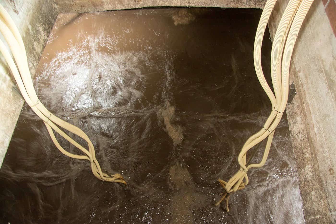 Draufsicht in ein betoniertes Klärbecken mit strömendem, braunem Abwasser. Von den Wänden führen mehrere beige Kabel- und Schlauchbündel in das Wasser.