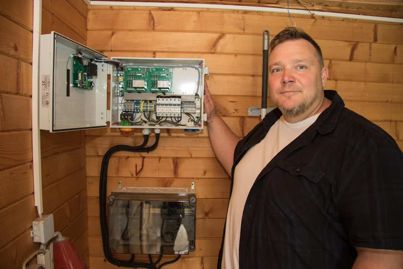Ein Mann steht in einem holzverkleideten Betriebsraum neben einem geöffneten Steuerungsgehäuse an der Wand, das elektronische Platinen und Anschlüsse zeigt.