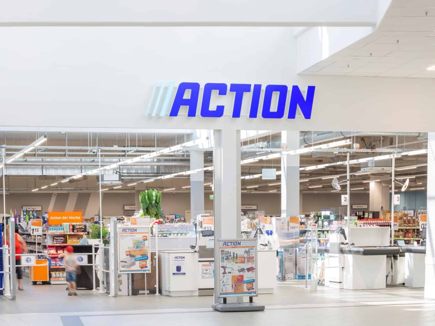 Blick auf den hellen Eingangsbereich einer Filiale der Handelskette Action mit blauem Schriftzug über dem Entree. Dahinter sind weiße Kassenbänder, Hinweisschilder und gefüllte Regale im Verkaufsraum der Mall sichtbar.