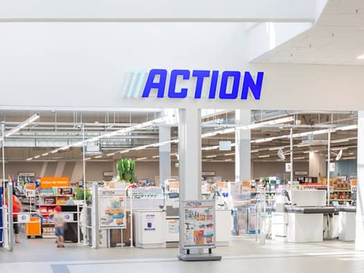 Blick auf den hellen Eingangsbereich einer Filiale der Handelskette Action mit blauem Schriftzug über dem Entree. Dahinter sind weiße Kassenbänder, Hinweisschilder und gefüllte Regale im Verkaufsraum der Mall sichtbar.