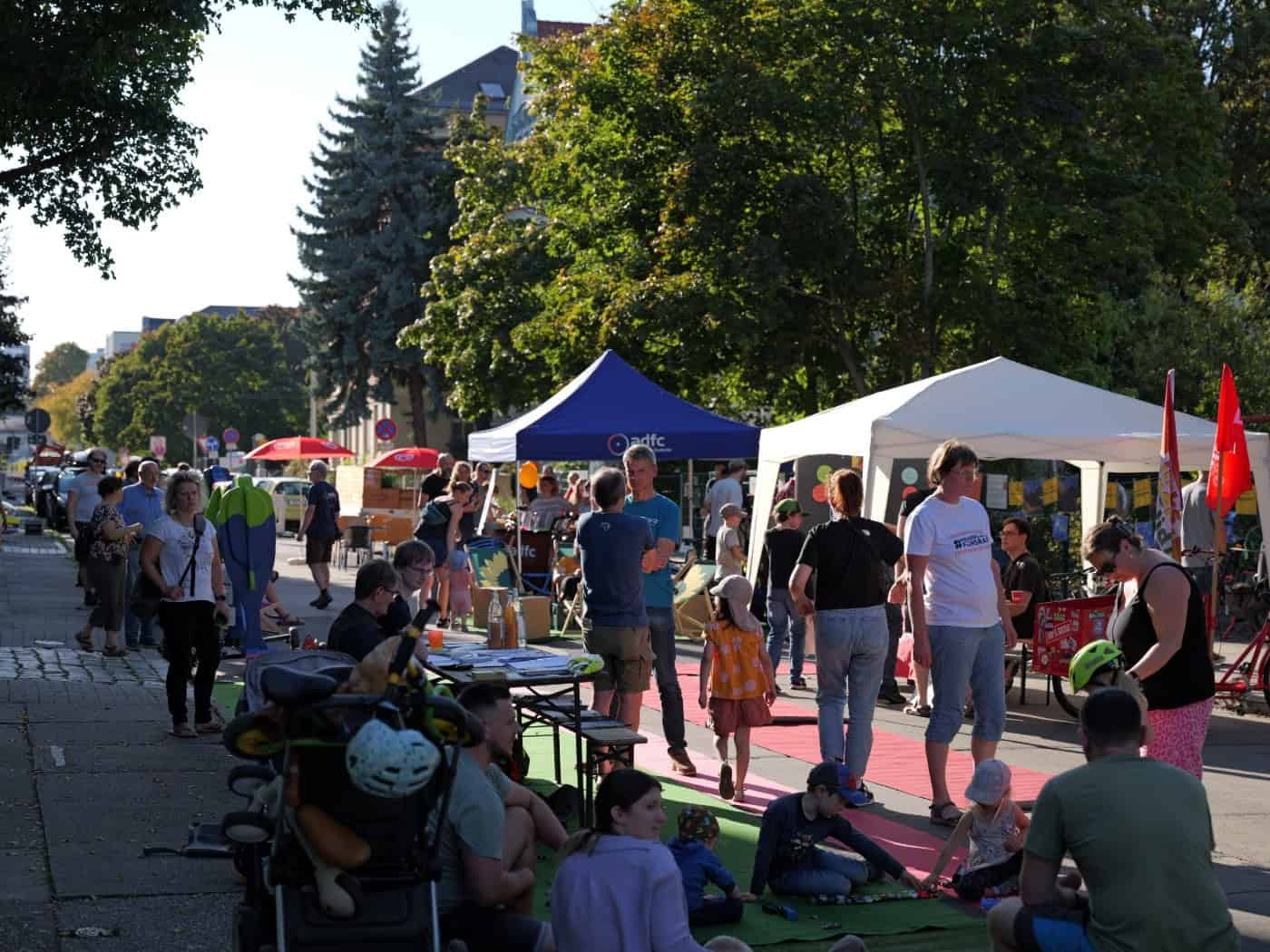 Belebte Straße während einer Aktion: Menschen stehen unter einem blauen ADFC-Pavillon und einem weißen Zelt zusammen, unterhalten sich und sitzen an Tischen. Im Vordergrund spielen Kinder auf einem ausgelegten Teppich auf der Straße.