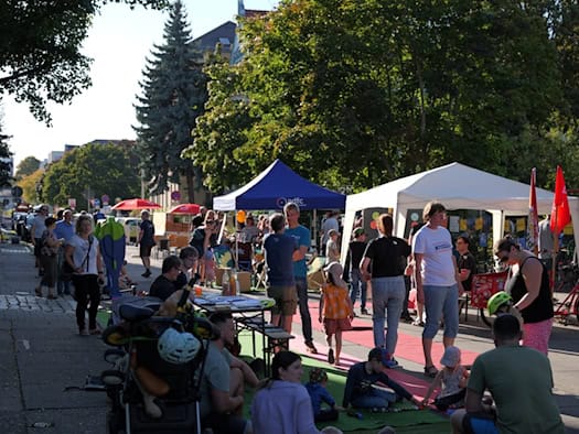 Belebte Straße während einer Aktion: Menschen stehen unter einem blauen ADFC-Pavillon und einem weißen Zelt zusammen, unterhalten sich und sitzen an Tischen. Im Vordergrund spielen Kinder auf einem ausgelegten Teppich auf der Straße.