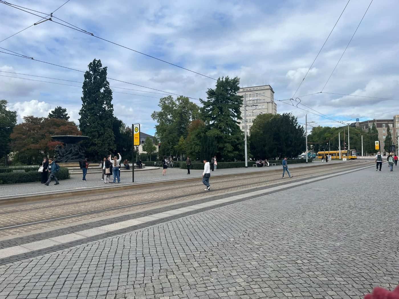 Weitwinkelaufnahme des Dresdner Albertplatzes bei leicht bewölktem Himmel. Im Vordergrund erstreckt sich gepflasterter Boden mit Straßenbahngleisen. Auf den Gehwegen warten Menschen an einer Haltestelle, im Hintergrund stehen grüne Bäume und ein großer Brunnen.