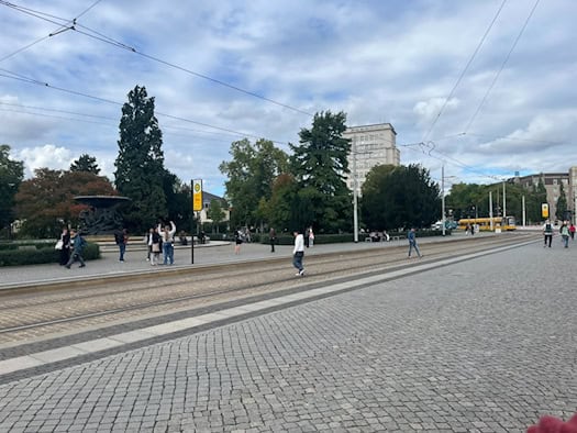 Weitwinkelaufnahme des Dresdner Albertplatzes bei leicht bewölktem Himmel. Im Vordergrund erstreckt sich gepflasterter Boden mit Straßenbahngleisen. Auf den Gehwegen warten Menschen an einer Haltestelle, im Hintergrund stehen grüne Bäume und ein großer Brunnen.