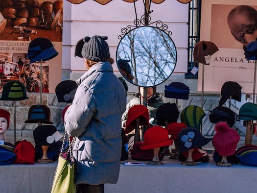 Eine Frau in grauer Winterjacke steht von hinten gesehen vor einem bunten Marktstand für handgefertigte Hüte. Auf dem Tisch stehen Hutständer mit Mützen und Hüten in Rot, Blau und Schwarz sowie ein runder Spiegel in der Mitte.