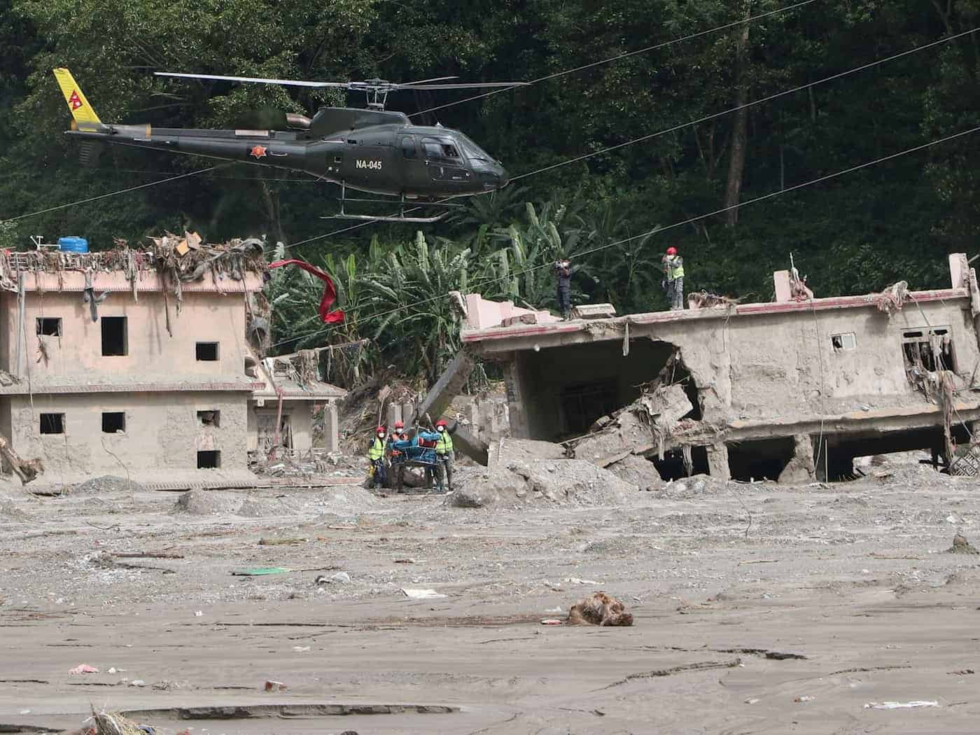 Ein olivgrüner Hubschrauber fliegt über durch ein Hochwasser schwer beschädigten, grauen Betonhäusern. Rettungskräfte mit Schutzhelmen stehen auf dem Dach eines zerstörten Gebäudes und im Schlamm. Dichter, grüner Wald bildet den Hintergrund.