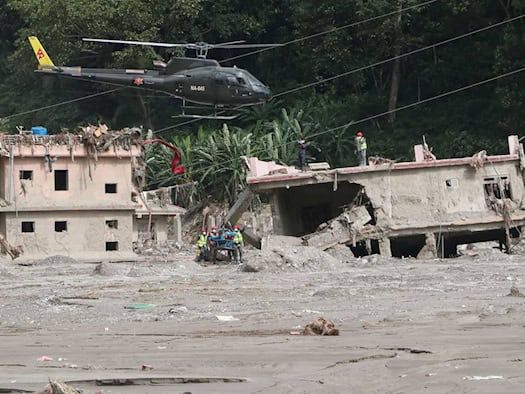 Ein olivgrüner Hubschrauber fliegt über durch ein Hochwasser schwer beschädigten, grauen Betonhäusern. Rettungskräfte mit Schutzhelmen stehen auf dem Dach eines zerstörten Gebäudes und im Schlamm. Dichter, grüner Wald bildet den Hintergrund.