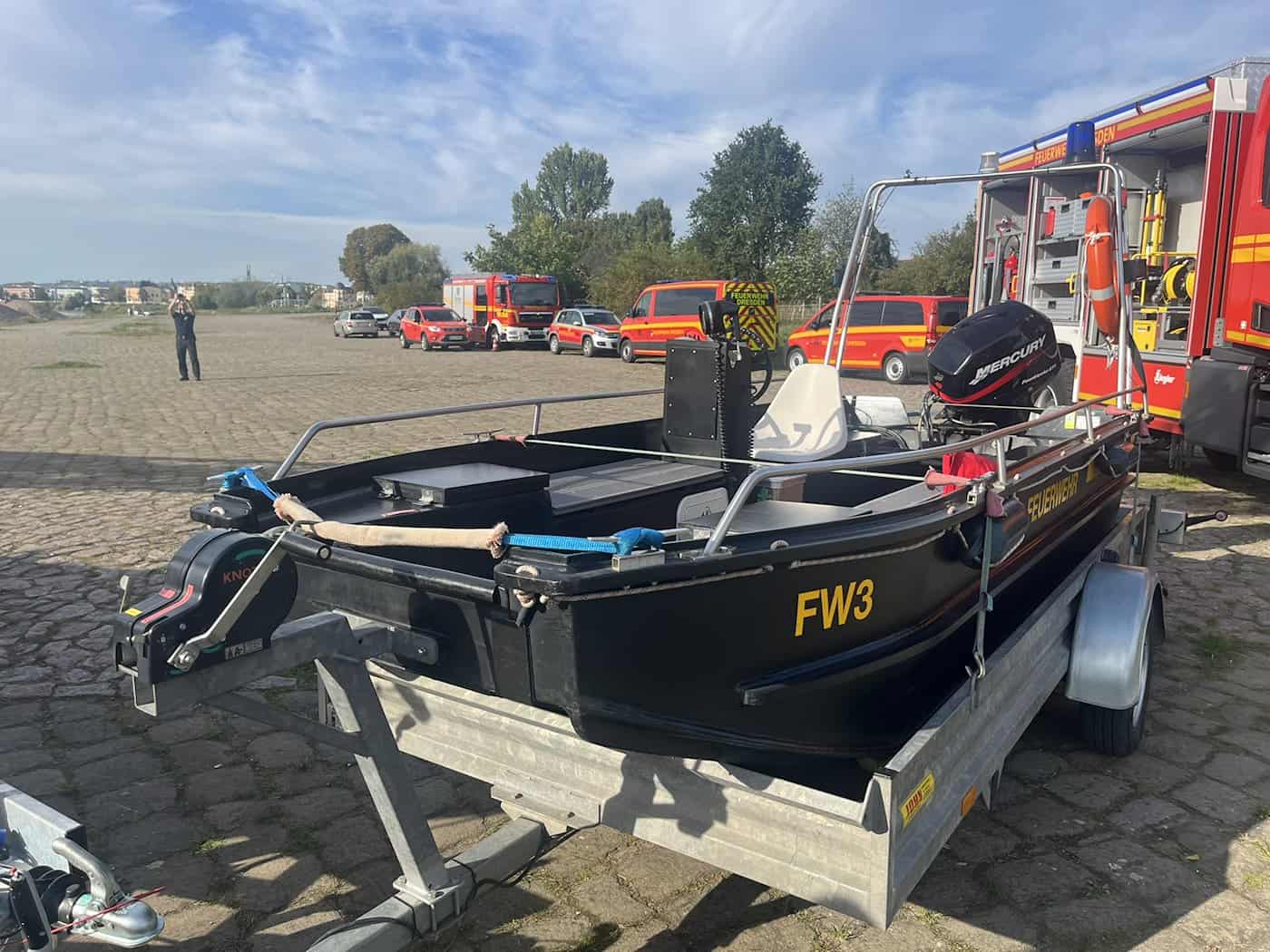 Nahaufnahme eines älteren, schwarzen Mehrzweckbootes der Feuerwehr Dresden mit Aufschrift FW3 auf einem Anhänger. Im Hintergrund stehen weitere Einsatzfahrzeuge auf dem Pflasterplatz.