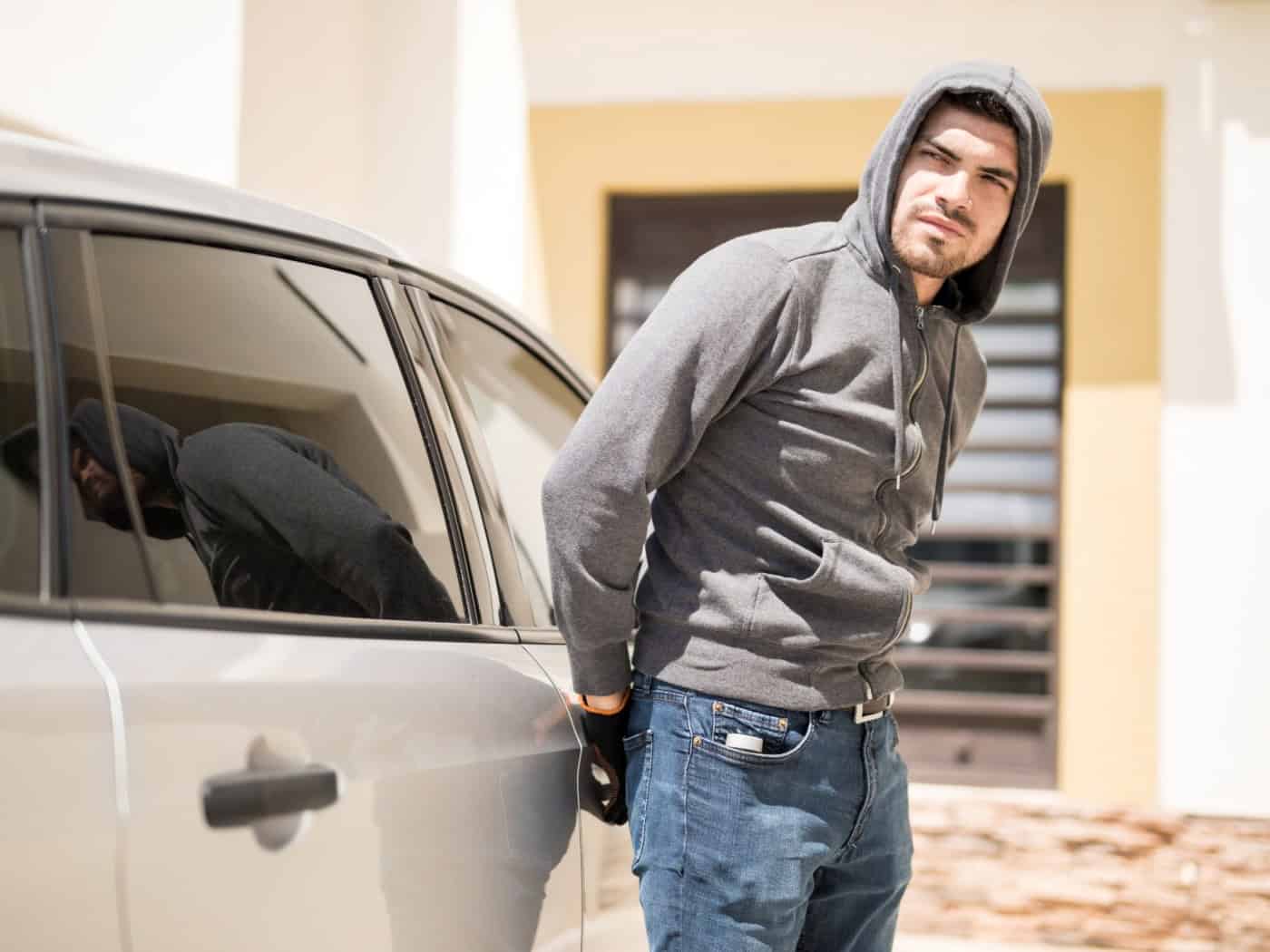 Symbolbild: Ein Mann in einem grauen Kapuzenpullover schaut sich vorsichtig um, während er versucht, die Tür eines silbernen Autos aufzubrechen.