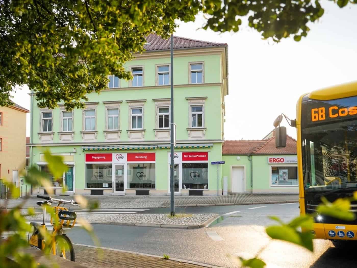 Außenansicht eines hellgrünen Gebäudes am Kreisverkehr mit AWO-Aushängen wie „Begegnung“ und „Beratung“ im Erdgeschoss. Im Vordergrund ein gelbes Leihfahrrad, rechts fährt ein gelber Dresdner Linienbus der Linie 68 vorbei.