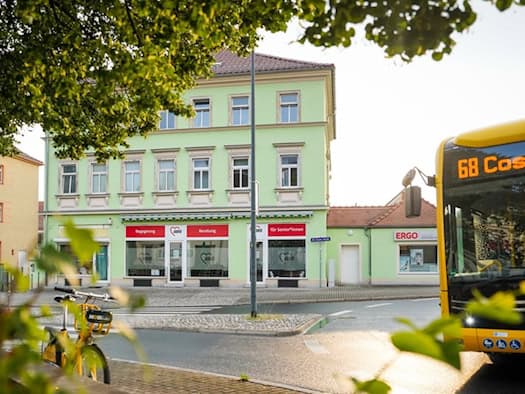 Außenansicht eines hellgrünen Gebäudes am Kreisverkehr mit AWO-Aushängen wie „Begegnung“ und „Beratung“ im Erdgeschoss. Im Vordergrund ein gelbes Leihfahrrad, rechts fährt ein gelber Dresdner Linienbus der Linie 68 vorbei.
