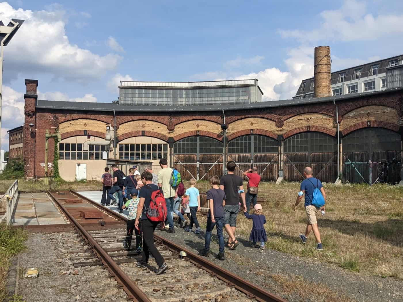 Eine Besuchergruppe geht auf den Gleisen einer Schiebebühnenanlage vor dem historischen Backstein-Ringlokschuppen des Bahnbetriebswerks Dresden-Altstadt. Im Hintergrund erstrecken sich die Tore des Lokschuppens und ein Schornstein unter leicht bewölktem Himmel.