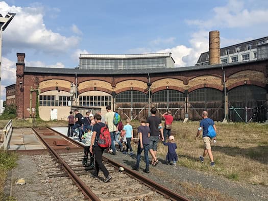 Eine Besuchergruppe geht auf den Gleisen einer Schiebebühnenanlage vor dem historischen Backstein-Ringlokschuppen des Bahnbetriebswerks Dresden-Altstadt. Im Hintergrund erstrecken sich die Tore des Lokschuppens und ein Schornstein unter leicht bewölktem Himmel.