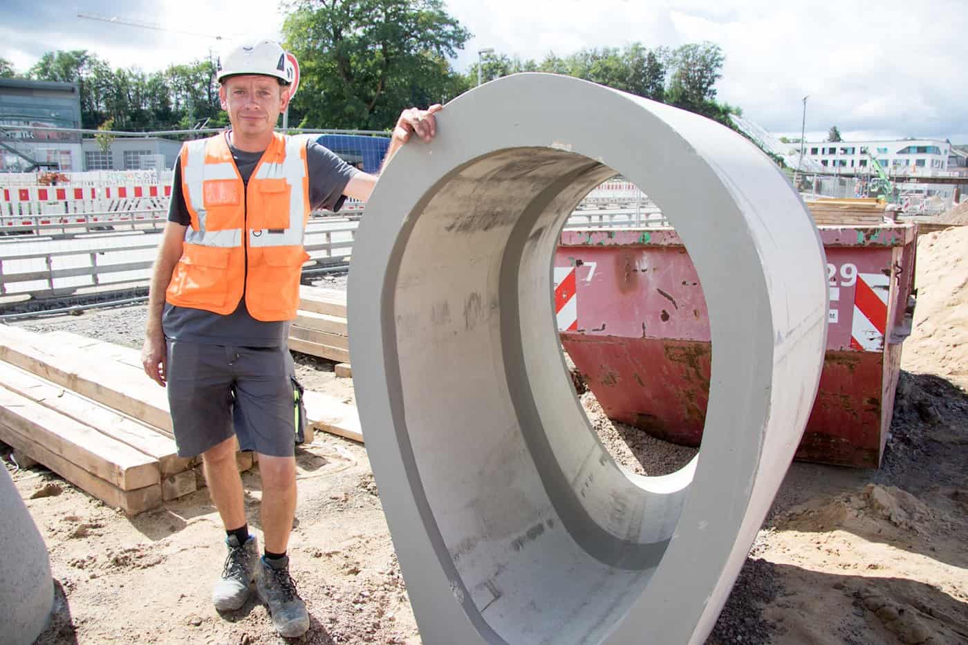 Ein Bauarbeiter in orangefarbener Warnweste und Helm stützt sich lächelnd auf ein großes ovales Eiprofil-Betonelement. Im Hintergrund stehen Bauschutt-Container und Absperrungen.