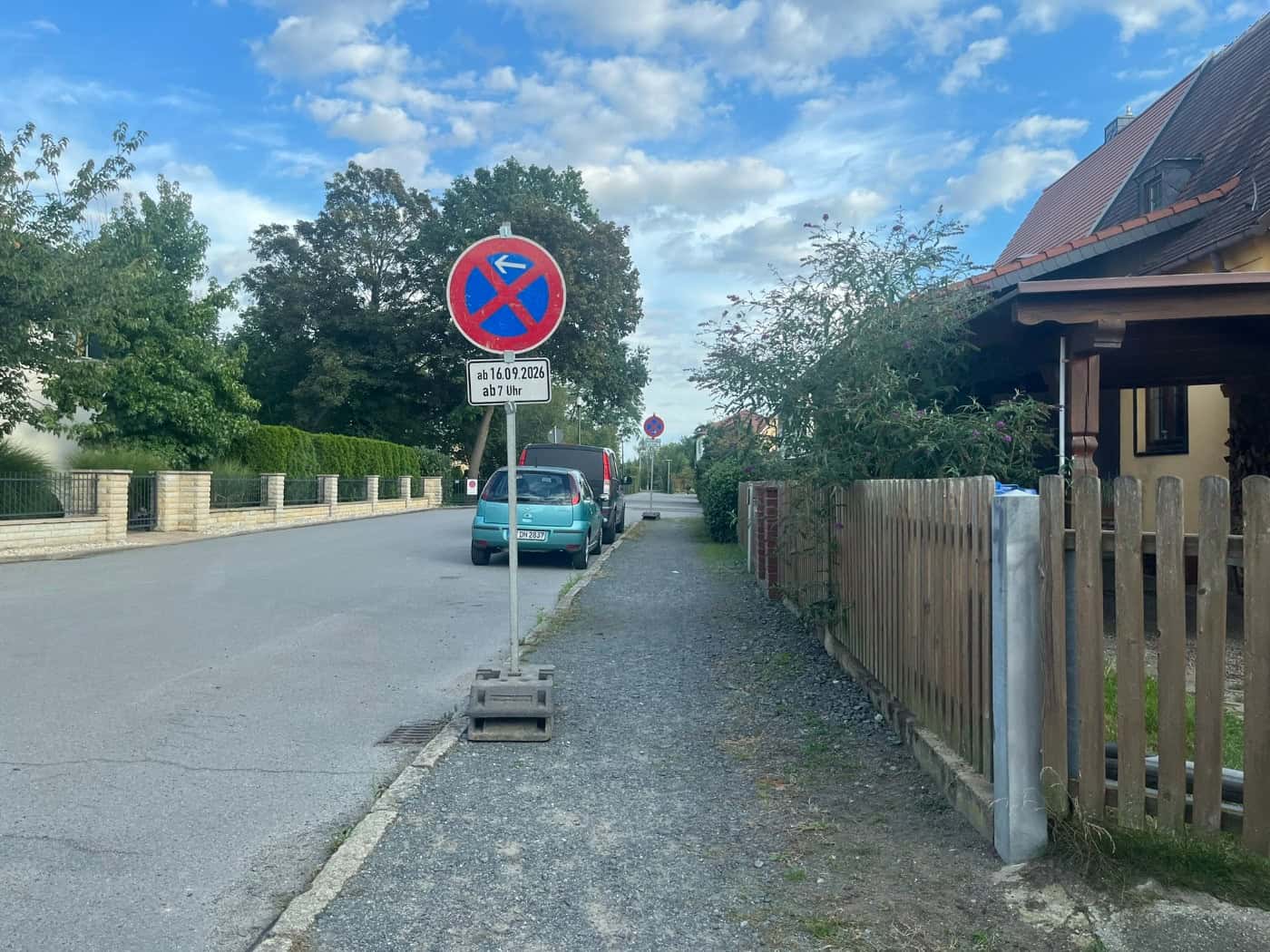 Ein mobiles Halteverbotsschild mit dem Zusatzschild „ab 16.09.2026 ab 7 Uhr“ steht am Gehweg einer Wohnstraße. Dahinter stehen geparkte Autos vor Einfamilienhäusern und Bäumen unter leicht bewölktem Himmel.