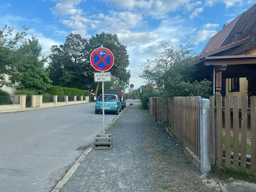 Ein mobiles Halteverbotsschild mit dem Zusatzschild „ab 16.09.2026 ab 7 Uhr“ steht am Gehweg einer Wohnstraße. Dahinter stehen geparkte Autos vor Einfamilienhäusern und Bäumen unter leicht bewölktem Himmel.