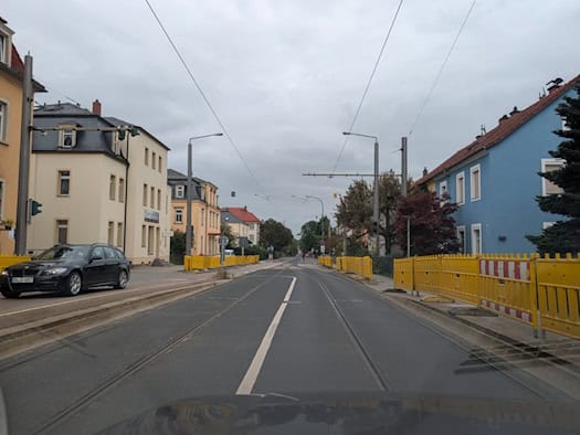 Blick aus einem Fahrzeug auf eine asphaltierte Straße mit Straßenbahngleisen in Dresden-Klotzsche. Gelbe Baustellen-Schutzzäune säumen die Gehwege links und rechts vor mehrgeschossigen Wohnhäusern unter bedecktem Himmel.